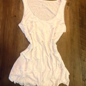 Vit kort klänning med volanger och strass - Supersöt vit kort klänning med volanger och strassdetaljer längs halsringningen. Klänningen är ärmlös och har en lätt, flowy passform som ger en chill vibe. Perfekt för varma dagar och har en feminin touch med de glittrande detaljerna.