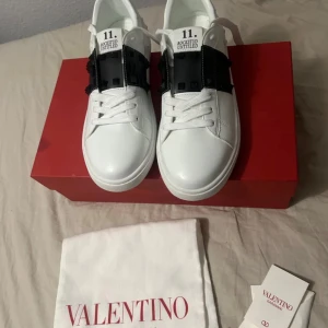 Valentino Rockstud Untitled sneakers - Snygga vita Valentino Rockstud Untitled sneakers med svart läderband och ikoniska pyramidnitar över vristen. Skorna har rund tå, platt sula och vita snören. Endast lådan och Skorna följer med