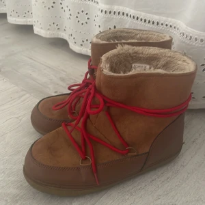 Chestnut boots med röd snörning! - Säljer ett par bruna moon boots med röd snörning och mjukt, beige foder på insidan. Skorna har platt sula och är tillverkade i syntetmaterial med mockaliknande yta. Perfekta för kalla vinterdagar och har en rund tå för extra komfort.
