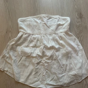 Vit bandeau tubtopp i linne - Säljer en ljusvit bandeau tubtopp med smockad rygg och lätt rynkad nederdel. Toppen är axelbandslös och har en flowy passform som ger en chill och somrig vibe. Materialet är linne, vilket gör den luftig och skön att bära under varma dagar.