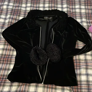 Svart sammet kavaj från Talace - Svart kavaj i mjuk sammet från Talace med snygga volangdetaljer vid krage och fickor. Jackan har en elegant siluett och dragkedja framtill. Perfekt för dig som gillar unika detaljer och vill sticka ut med din stil.