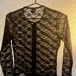 Svart spetsblus med knappar - Säljer en transparent svart blus i spets med lång ärm och knappar framtill. Översta knappen är en gullig liten rosa ros. Blusen har ett blommigt spetsmönster och rund halsringning. Nypris 350kr, aldrig använd.