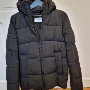 Svart pufferjacka från Calvin Klein - Svart pufferjacka från Calvin Klein i nyskick! Storlek:M Med huva och vadderad design. Jackan har dragkedja framtill och på fickorna, samt en snygg logotyp på ärmen. Tillverkad av 100% återvunnen polyester, både skal och foder. Perfekt för kalla dagar! Nypris:3000 
