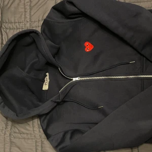 Svart hoodie från Comme des Garçons Play - Svart hoodie med dragkedja från Comme des Garçons Play. Tröjan har huva med snörning och ett broderat rött hjärta med ögon på bröstet. Tillverkad i mjuk bomull, perfekt för dig som gillar stilrena detaljer och streetwear vibes.