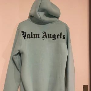 Ljusblå Palm Angels hoodie - Säljer en ljusblå hoodie från Palm Angels med stor svart logga på ryggen och mindre logga framtill. Tröjan har huva med snörning och en stor magficka. Perfekt för dig som gillar streetwear och vill sticka ut med en snygg färg.