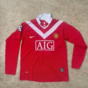 Manchester United långärmad Nike tröja - Röd Manchester United fotbollströja från Nike med lång ärm, vit V-mönster över bröstet och vit krage. Broderat klubbmärke, Premier League-patch på ärmen och AIG sponsortryck på framsidan. Tillverkad i lätt polyester med NikeFIT-teknologi.