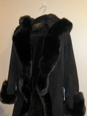 Svart pälsjacka från Giorgia Fashion XS - Säljer en svart pälsjacka från Giorgia Fashion i storlek XS. Jackan har fluffiga pälsdetaljer längs krage, ärmslut och huva, samt bälte i midjan och dragkedjor vid fickorna. Materialet är mjukt och ger en lyxig vibe, perfekt för kalla dagar. Använd 2 ggr. 
