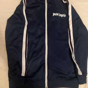 Marinblå Palm Angels zip hoodie M - Snygg marinblå zip hoodie från Palm Angels med vita ränder längs ärmarna och logga på bröstet. Tröjan har dragkedja framtill och ribbade muddar. Perfekt för dig som gillar streetwear och vill sticka ut med en ikonisk look.