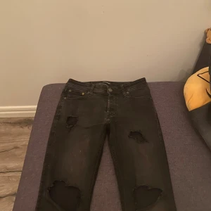 Svarta ripped jeans Jack & Jones - Svarta jeans från Jack & Jones med slitna detaljer och hål på benen för en edgy look. Jeansen har slim passform. Klassisk femficksmodell med dragkedja och knapp. Jeansen är i stretchigt denim som sitter snyggt och bekvämt. Jeansen är storlek 28/30