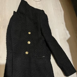 Svart kavaj med guldknappar från Zara - Snygg svart kavaj från Zara med sex guldiga knappar framtill. Jackan har en strukturerad, lite grov yta och klassisk krage. Passar dig som gillar stilrena och trendiga detaljer. Perfekt att styla med jeans eller kjol för en cool look.