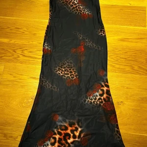 Svart långklänning med leopardmönster - Snygg svart långklänning med leopardmönster och röda rosor. Klänningen har en figurnära passform och är ärmlös, vilket ger en cool och trendig look. Materialet känns lätt och följsamt mot kroppen. Perfekt för dig som vill sticka ut med ett djuriskt och romantiskt print.
