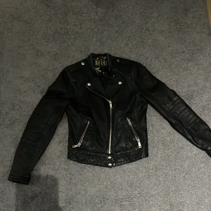 Svart skinnjacka Jofama Marie Serneholt - Snygg svart skinnjacka från Jofama by Marie Serneholt i storlek 36. Jackan har coola detaljer som silverfärgade dragkedjor, knappar och quiltade partier på axlarna. Fodret har ett blommigt mönster i guld. Perfekt för dig som vill ha en edgy look.