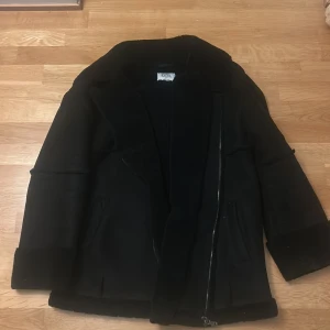 Svart teddyjacka med dragkedja - Svart teddyjacka från KARO KAUER med mjuk insida och bred krage. Jackan har två sidofickor, grova muddar och stängs med en rejäl dragkedja. Perfekt för kalla dagar och har en clean, oversized look som är enkel att styla.