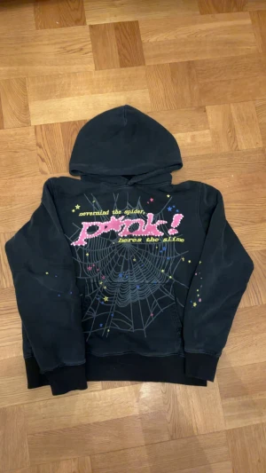 Sp5der hoodie - Cool svart hoodie från Sp5der med färgglatt tryck framtill som inkluderar text, stjärnor och ett spindelnätsmotiv. Tröjan har huva och ribbade muddar. Perfekt för dig som gillar unika och lekfulla plagg.