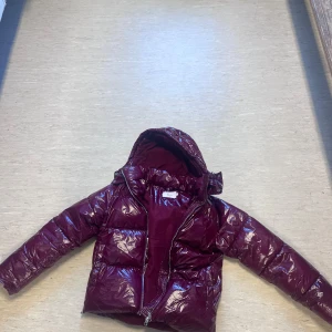 Vinröd pufferjacka med huva - Säljer en vinröd pufferjacka från Nelly. Knapparna i fickan är sönder men den funkar fortfarande. Den vinröd jackan  går inte att köpa längre.köpt för 800 hundra. Använt 10 gånger typ  med glansig yta och huva. Jackan har dragkedja framtill och är fodrad med mjukt tyg på insidan. Perfekt för kalla dagar och har en snygg, modern look som sticker ut. 