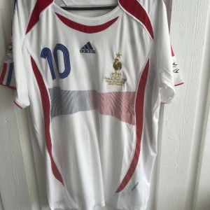Frankrike Zidane VM 2006 fotbollströja - Säljer en officiell Frankrike fotbollströja från VM 2006 med Zidane och nummer 10 på ryggen. Tröjan är vit med röda och blå detaljer, Adidas-logga på bröstet och broderat VM-märke. Tillverkad i lätt polyester med korta ärmar och rund hals.