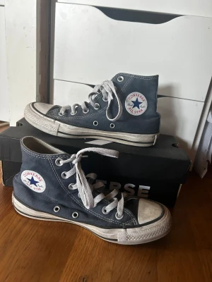 Converse skor - Säljer ett par klassiska Converse All Star high-top sneakers i mörkblå canvas med vit gummisula och rund tå. Skorna har vit snörning och den ikoniska logotypen på sidan. Extra vita skosnören medföljer. Perfekt för dig som gillar retrostil och streetwear.