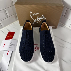Mörkblå sneakers från Christian Louboutin - Snygga mörkblå sneakers från Christian Louboutin med mockaöverdel och coola svarta nitar på tån. Skorna har snörning, vit platt sula och den ikoniska röda undersidan. Insidan är lyxigt fodrad i beige skinn. Perfekt för dig som vill sticka ut med exklusiv stil.
