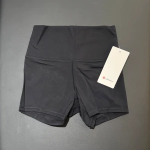 Svarta träningsshorts från Lululemon i storlek 4/S - Snygga svarta träningsshorts från Lululemon med hög midja och tight passform. Tillverkade i ett stretchigt material med 77% nylon och 23% spandex som ger grym komfort och rörelsefrihet. Perfekta för gymmet eller löprundan, med stilren design och diskret logga bak.