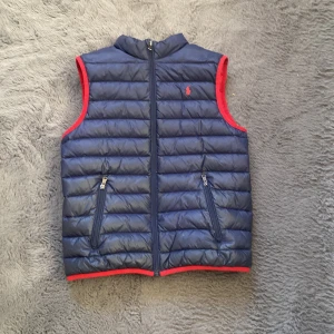 Mörkblå dunväst från Ralph Lauren - Snygg mörkblå dunväst från Ralph Lauren med röd insida och röda detaljer vid ärmhål och nederkant. Västen har quiltad design, två fickor med dragkedja och klassisk Polo-logga broderad i rött på bröstet. Västen dragkedjor kan vara lite tröga ibland.