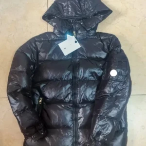 Svart Moncler pufferjacka med huva - Svart pufferjacka från Moncler med glansig finish, huva och klassisk Moncler-logga på ärmen. Jackan har två fickor med dragkedja och quiltad design. Insidan har en unik serietidningsetikett och autenticitetsmärkning med QR-kod. Perfekt för kalla dagar. Skriv för mer info! 