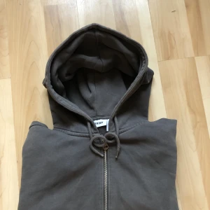 Mörkgrå hoodie från Weekday - Säljer en mörkgrå hoodie från Weekday med dragkedja framtill och snörning i huvan. Tröjan har långa ärmar, ribbade muddar och en stor ficka på magen. Perfekt för chill dagar och enkel att matcha med jeans eller joggers.