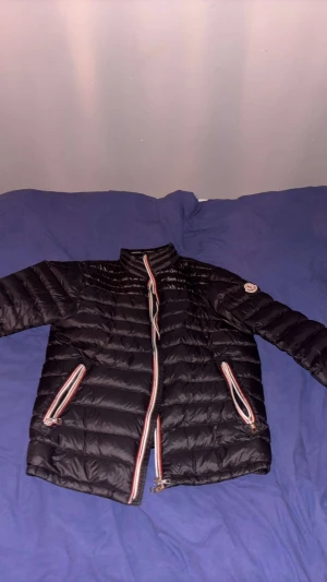 Svart Moncler pufferjacka med dragkedja - Svart pufferjacka från Moncler med klassisk quiltad design och coola rödvita detaljer på dragkedjorna. Jackan har hög krage, två fickor med dragkedja och Moncler-logga på ärmen. Perfekt för dig som vill ha en snygg och varm jacka med streetkänsla.