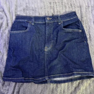 Mörkblå jeanskjol med fickor - Snygg mörkblå jeanskjol med klassiska fickor både fram och bak samt hällor för bälte. Kjolen har kontrastsömmar och en rak, kort passform som ger en cool och avslappnad vibe. Perfekt att matcha med sneakers eller boots för en trendig look.