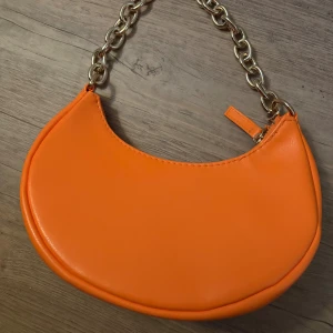Orange axelväska från H&M med kedja - Cool orange axelväska från H&M med chunky guldfärgad kedja som axelrem. Väskan har en mjukt rundad form och stängs med dragkedja upptill. Tillverkad i syntetmaterial och perfekt för att addera färg till din outfit.
