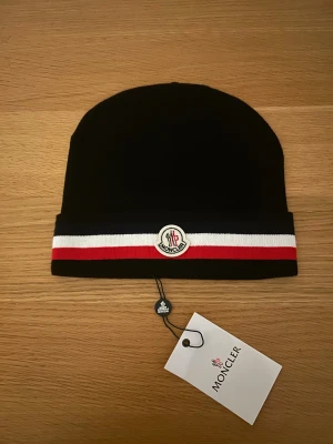 Svart mössa med ränder - Svart mössa med klassisk logga framtill. Mössan har en bred kant med randigt mönster i vitt, rött och mörkblått. Snygg och stilren design som lyfter din look, perfekt för kalla dagar. Materialet är mjukt och stickat för extra komfort.