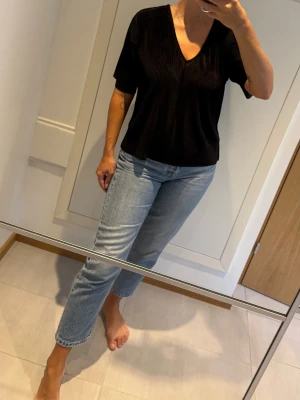 Svart plisserad blus med v-ringning - Säljer en svart plisserad blus med v-ringad hals och korta ärmar. Blusen har en luftig passform och är tillverkad i ett lätt, glansigt syntetmaterial som ger en snygg struktur. Svart ribbad skir v-ringad kortärmad topp. Perfekt till tex julfest och nyår.