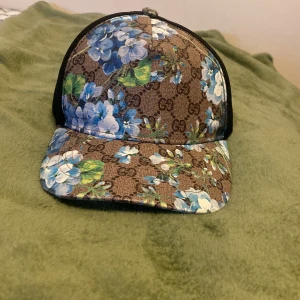 Gucci blommig keps med mesh - Snygg keps från Gucci med brunt GG-mönster och blommigt print i blått, vitt och grönt. Framsidan är i canvas med blommor, baksidan i svart mesh och justerbart band med klassiska Gucci-ränder. Perfekt för dig som vill sticka ut med designerstil.