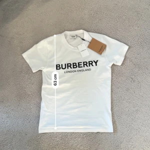 Vit Burberry t-shirt med logga - Vit t-shirt från Burberry med klassisk svart logotyp 'BURBERRY LONDON ENGLAND' tryckt på bröstet. Rund halsringning och korta ärmar. Tillverkad i mjuk bomull för en skön känsla. Perfekt för dig som gillar stilrena designer och ikoniska märken.Aktiv 24/7✅ Skickas inom 24h📦🚚 Kan gå ner i hugget 😃✅ Hör av er via frågor⁉️