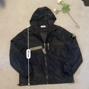 Svart vindjacka Stone Island L - Svart vindjacka från Stone Island med huva och två snedställda dragkedjefickor framtill. Jackan har den klassiska Stone Island-loggan på vänster ärm och är tillverkad i lätt polyester. Perfekt för dig som gillar streetwear och vill ha en clean look. Perfekt för hösten och vintern. Aktiv 24/7✅ Skickas inom 24h📦🚚 Kan gå ner i hugget 😃✅ Hör av er via frågor⁉️