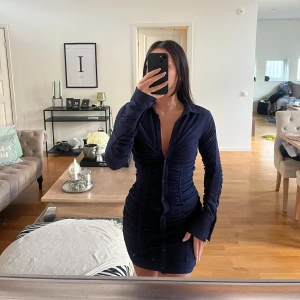 Marinblå klänning från Zara - Säljer en marinblå kort skjortklänning med lång ärm och djup v-ringning. Klänningen har knappar hela vägen framtill och är figurnära med snygga rynkade detaljer längs sidorna. Perfekt för dig som gillar en stilren och modern look.