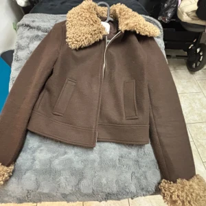Brun teddyjacka från Stradivarius - Säljer en brun teddyjacka från Stradivarius i storlek XS. Jackan har fluffig teddykrage och muddar i beige, två fickor framtill och stängs med dragkedja. Croppad modell som ger en cool och trendig look, perfekt för höst och vinter.