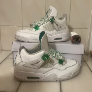 Air Jordan 4 Retro metallic green  - Nike Air Jordan 4 Retro sneakers i vitt och grönt, med coola gröna detaljer och mönstrade snören. Skorna har en klassisk meshpanel på sidan, synlig Air-enhet i sulan och Jumpman-logga på tungan och undersidan. Tillverkade i skinn och syntetmaterial för en schysst look. skosnören och crep skydd kommer med skorna.