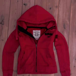 Röd hoodie från Diesel med dragkedja - Säljer en röd hoodie från Diesel i storlek small. Tröjan har huva med svarta broderade bokstäver, hel dragkedja framtill och två fickor. Materialet är mjukt och känns som bomull, perfekt för chill dagar. Diesel-logga på insidan och cool retro/archive-vibe.