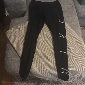 Nike Dri-Fit svarta leggings M - Svarta Nike Dri-Fit leggings med vit randig logga längs benet. Tight passform och hög midja, tillverkade i syntetmaterial som andas och transporterar bort fukt. Perfekta för träning och sportiga aktiviteter.