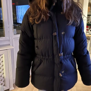 Mörkblå  dunjacka från Boomerang S - Mörkblå  dunjacka från Boomerang, modell Alexandra  med huva , dragkedja och snygga knappar framtill. Jackan har en puffad look, dragsko i midjan och två stora fickor. Perfekt för kalla dagar och har en klassisk stil som funkar till det mesta.. Den är precis kemtvättad och har också fixat dragkedjan, men den är fortfarande lite trög men fungerar absolut. En liten reva( 1cm) på ena ärmens baksida. Tjock och fluffig dunjacka som man aldrig fryser med. Storlek S men funkar även om man har XS .