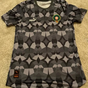 Marocko fotbollströja Puma svart/grå - Snygg Marocko fotbollströja från Puma med ett unikt geometriskt mönster i svart och grått. Kortärmad modell med rund hals och tryckt emblem på bröstet. Materialet är lätt och andas, perfekt för match eller träning. Arabisk text på ryggen ger extra stil.