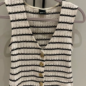 Randig stickad väst från Gina Tricot - Säljer en stickad väst från Gina Tricot i storlek XS. Västen är vit med svarta ränder, har v-ringning och fyra snygga knappar framtill. Perfekt att styla över en t-shirt eller skjorta för en cool look.