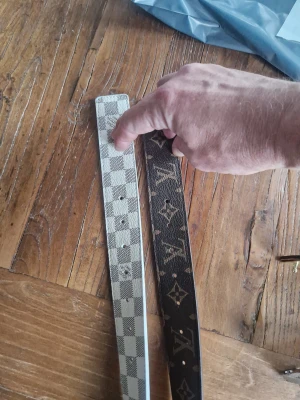 Louis Vuitton bälten i skinn110 - Två snygga Louis Vuitton bälten i skinn. Ett är rutigt i vitt och grått med klassiskt Damier-mönster, det andra är svart med det ikoniska LV-monogrammet i guld. Perfekta accessoarer för att lyfta din outfit med lyxig känsla.