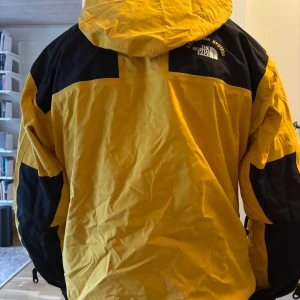 Gul och svart skidjacka från The North Face - Säljer vintage skidjacka från The North Face Expedition System. Jackan har huva, snörning i midjan, dragkedja och knappar framtill. Flera praktiska fickor med dragkedja och cool broderad logga på bröstet. Perfekt för dig som gillar street och outdoor.