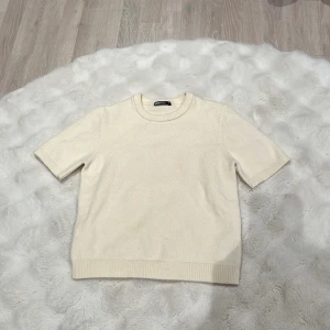 Krämvit stickad tröja från Gina Tricot - En enkel och stilren krämvit stickad tröja från Gina Tricot med korta ärmar och rund hals. Tröjan har ribbade muddar vid ärmslut och nederkant, vilket ger en snygg och clean look. Perfekt för lager-på-lager under kyliga dagar.