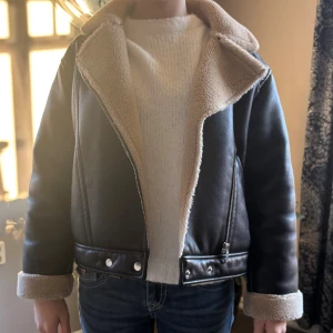 Svart teddyjacka med beige foder - Säljer en svart teddyjacka med mjukt beige foder och bred krage. Jackan har dragkedja framtill, detaljer med knappar vid midjan och uppvikta ärmslut med teddy. Snygg och varm modell som ger en cool vibe till din outfit.