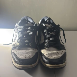 Nike Dunk Low svart/vit sneakers - Nike Dunk Low sneakers i svart och vitt med klassisk Swoosh på sidan. Skorna har snörning, rund tå och platt sula. Ovandelen är i skinn med perforerade detaljer på tån för extra ventilation. Perfekt för dig som gillar retrostil och streetwear.