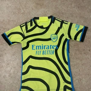 Arsenal gul och svart matchtröja - Säljer en Arsenal fotbollströja från Adidas i neon-gul med svarta och blå detaljer. Tröjan har korta ärmar och ett unikt prickigt och randigt mönster. Materialet är lätt och ventilerande polyester, perfekt för träning eller match. Tryck med Emirates Fly Better på bröstet.