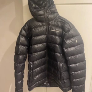 Svart Peak Performance dunjacka XL - Svart dunjacka från Peak Performance i storlek XL. Jackan har huva, dragkedja framtill och diskret logga på bröstet. Den är quiltad och har en clean, sportig look som funkar perfekt när det är kallt ute.