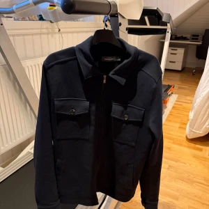 Mörkblå overshirt från Dressmann - Snygg mörkblå overshirt från Dressmann med två stora bröstfickor med knapp, klassisk krage och dragkedja framtill. Färg, mörk blå storlek M, köpt för 800 kr